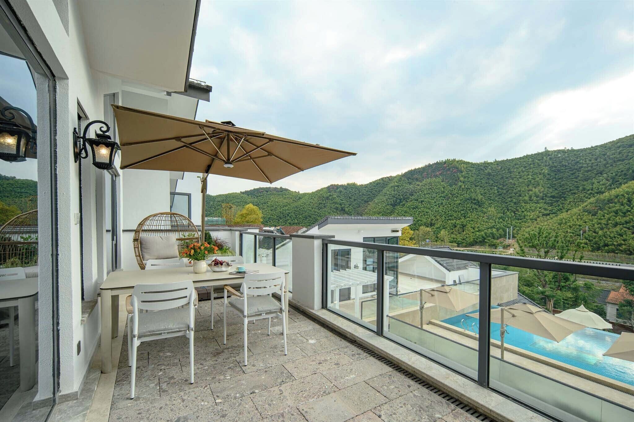 Un balcón con una mesa y sillas, un gran paraguas y vistas a una piscina y colinas verdes.