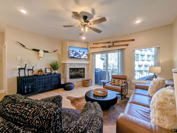 A living room with a fireplace, a ceiling fan, and a large window. - New Luxury | Keystone Lake | King Bed, Shuttle (Keystone)