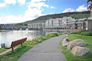 A lakeside walkway with a bench, large rocks, and a row of multi-story buildings with green roofs. - New Luxury | Keystone Lake | King Bed, Shuttle (Keystone)