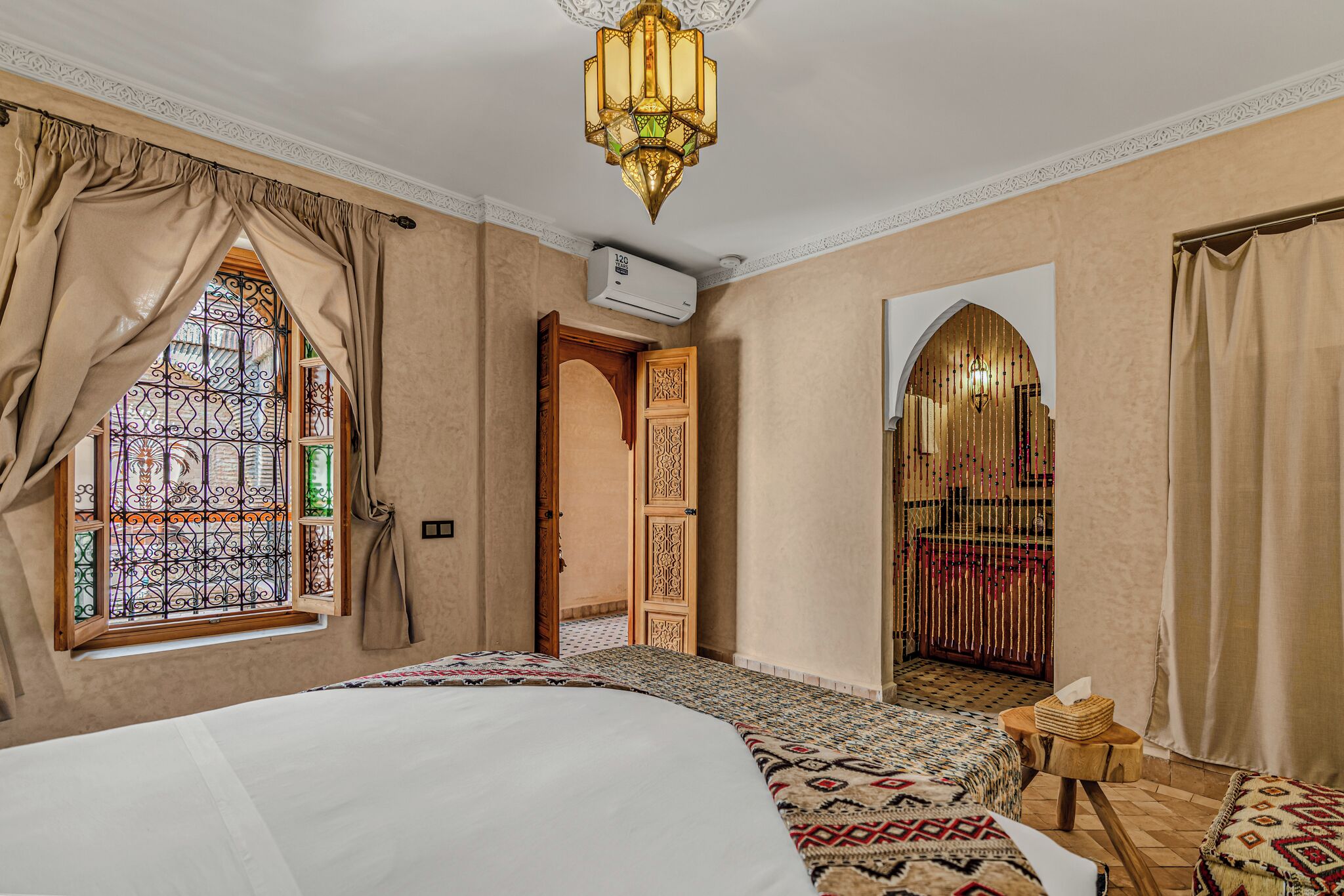 A hotel room with a large bed, a chandelier, a window with stained glass, and a door leading to another room.
