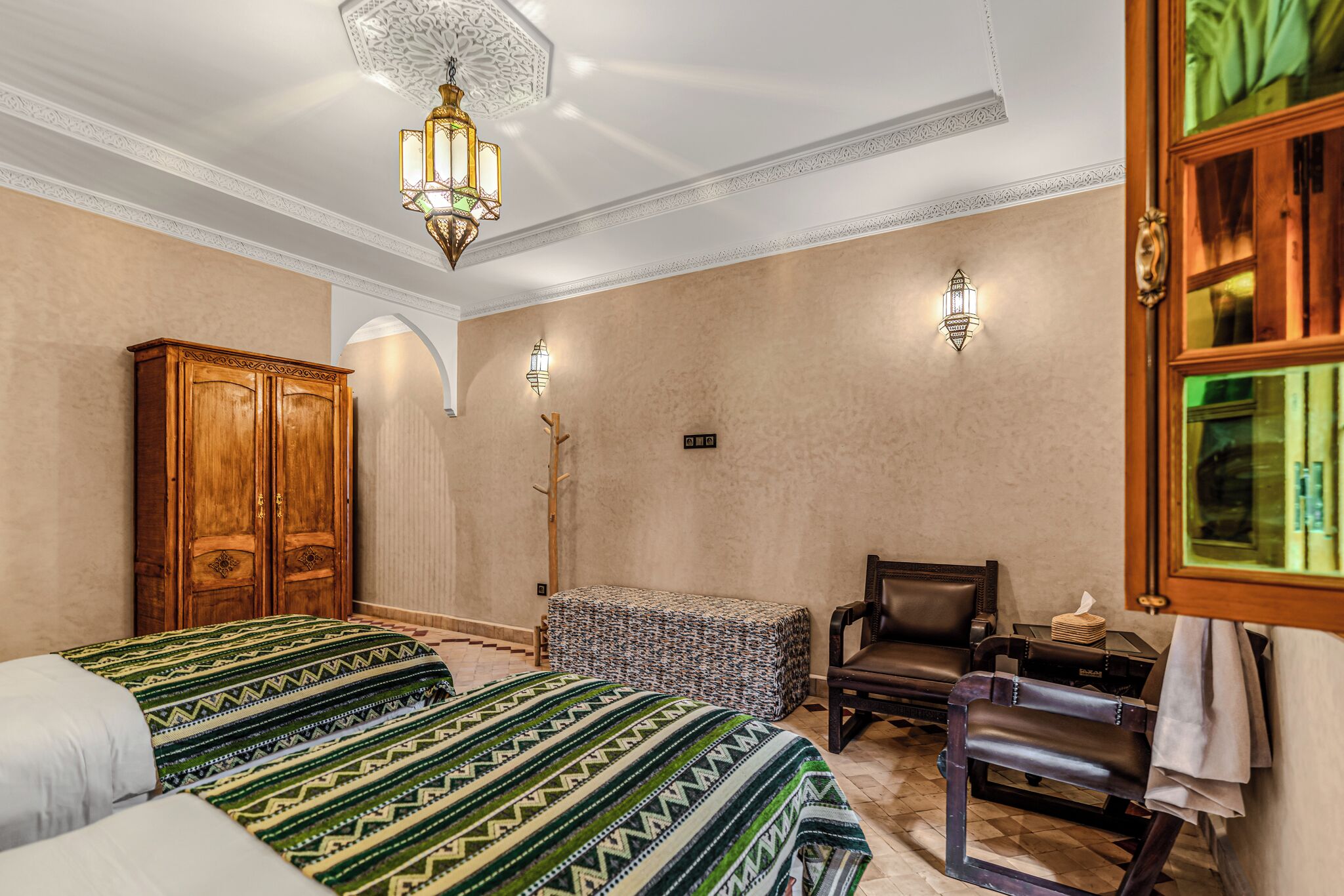 Quarto de hotel com duas camas, um guarda-roupa de madeira, uma cadeira, uma mesinha e uma luminária decorativa no teto.