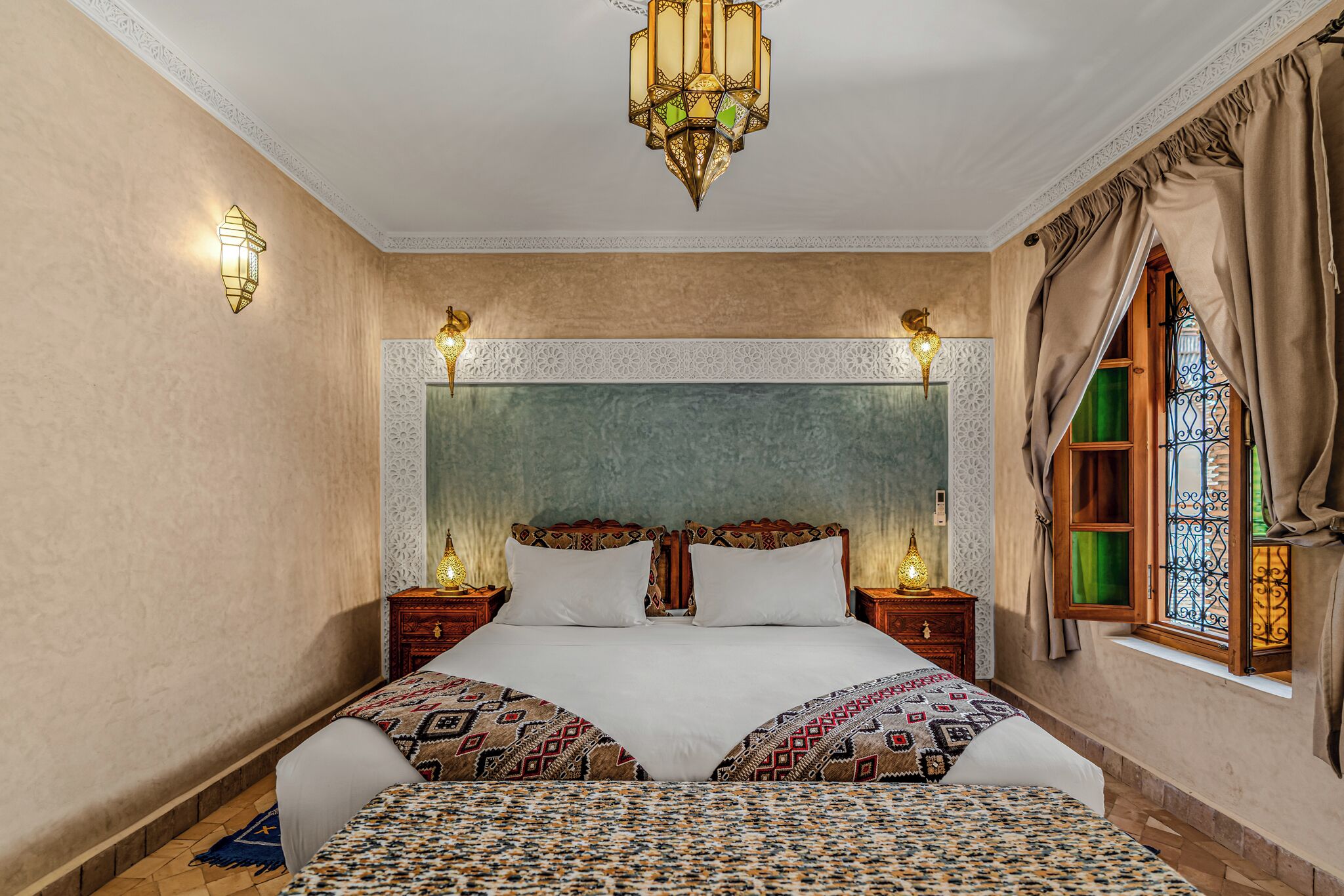 Un dormitorio con una cama grande, dos mesitas de noche con lámparas, una lámpara decorativa en el techo y una ventana con cortinas.