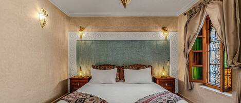 A bedroom with a large bed, two bedside tables with lamps, a decorative ceiling light, and a window with curtains.