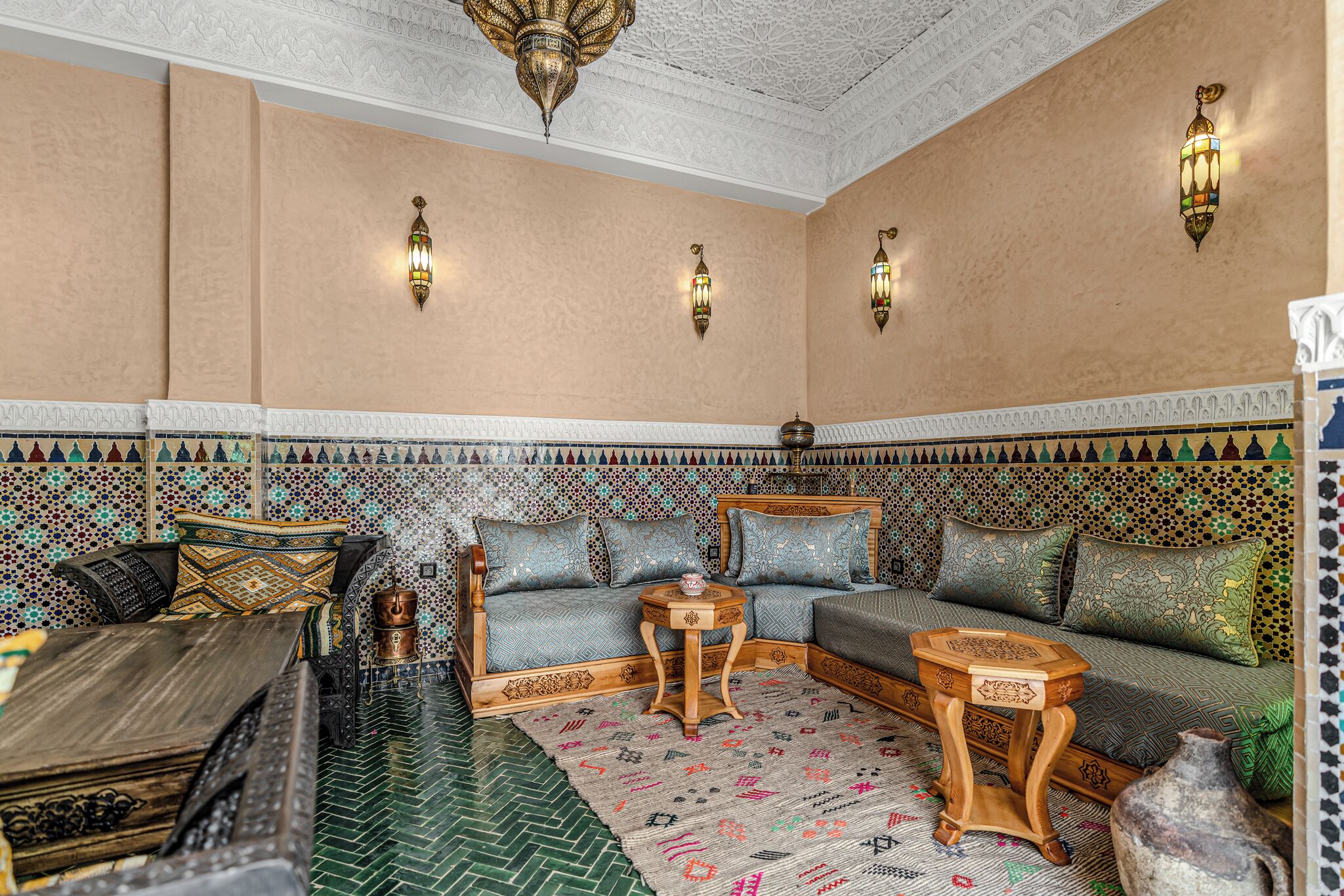 Een traditioneel Marokkaans interieur met een patroonmuur, een zithoek, een houten bijzettafeltje en een decoratieve lantaarn.