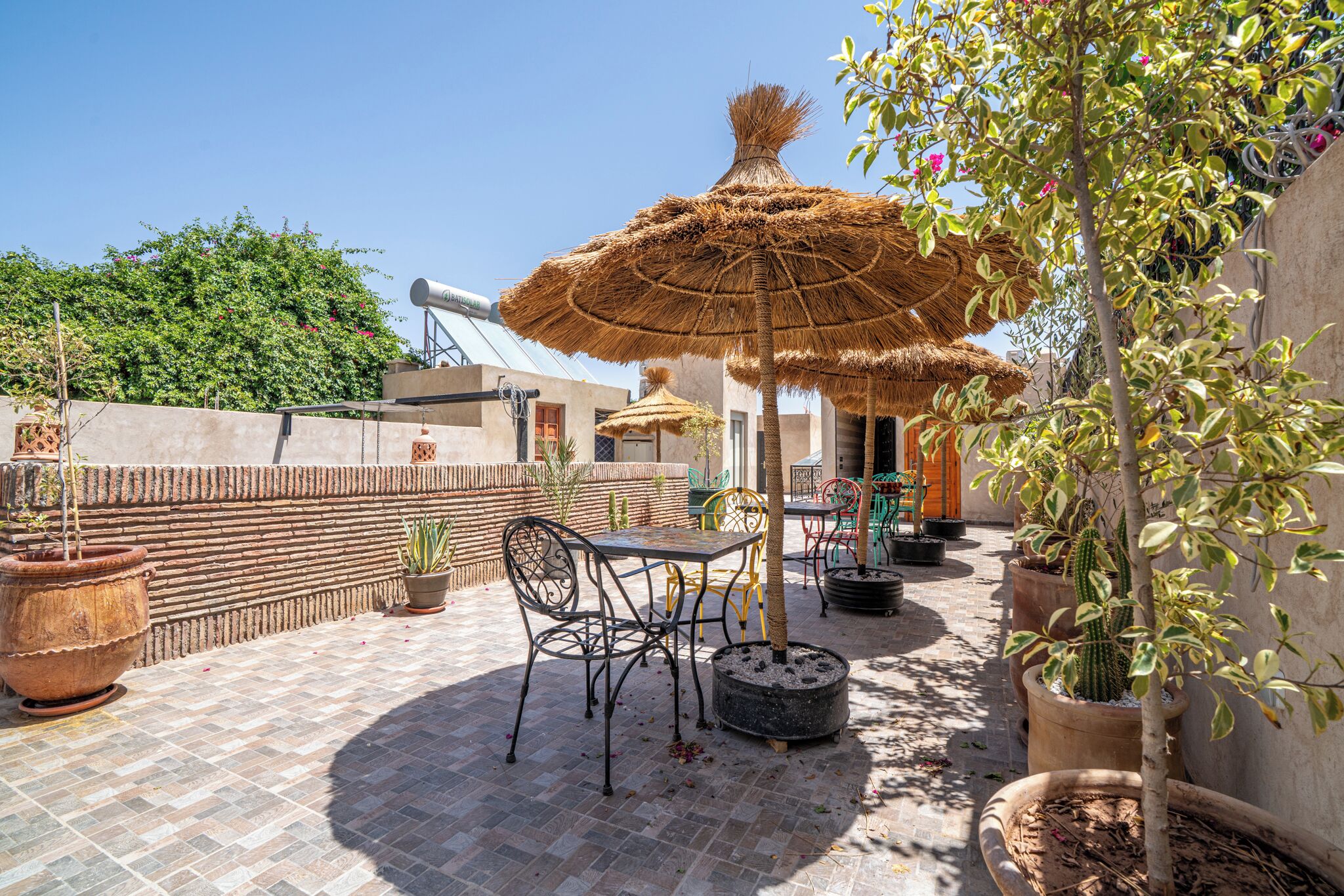Un patio al aire libre con una mesa y sillas, rodeado de plantas en macetas y un paraguas de paja.