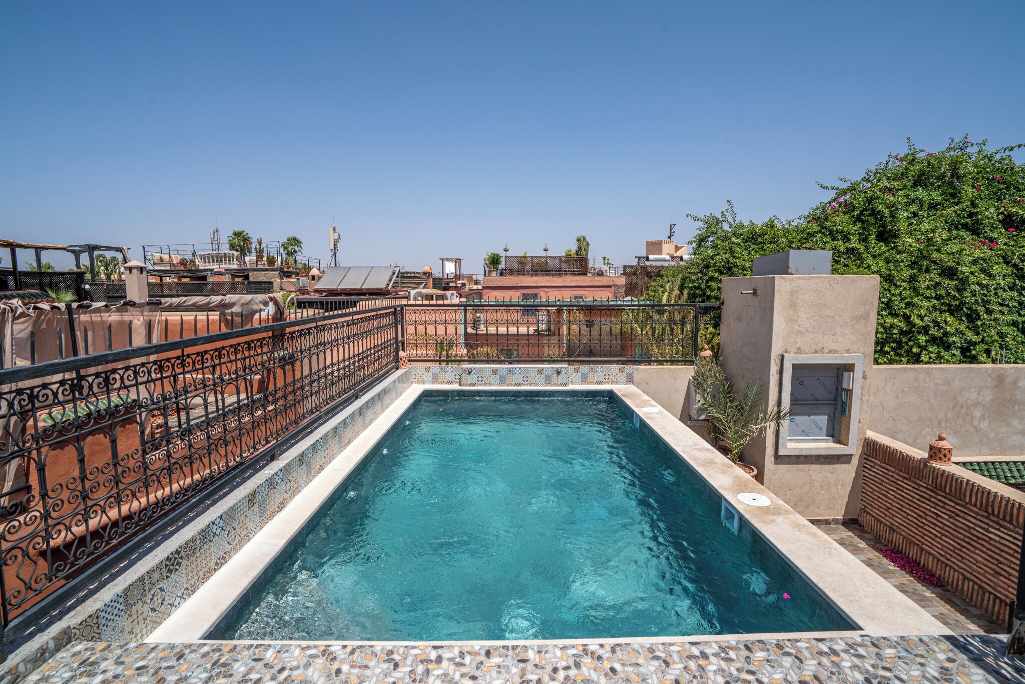 Piscina rectangular con bordeado de azulejos, rodeada por una valla metálica decorativa, con edificios y follaje de fondo.