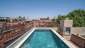 A rectangular swimming pool with a tiled border, surrounded by a decorative metal fence, set against a backdrop of buildings and greenery. - Riad Maison D'HÔTE ABDEL & JAMILA (Marrakech)