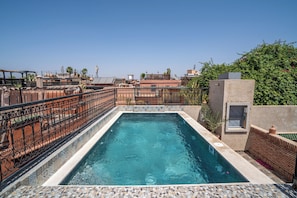 Piscina rectangular con bordeado de azulejos, rodeada por una valla metálica decorativa, con edificios y follaje de fondo.