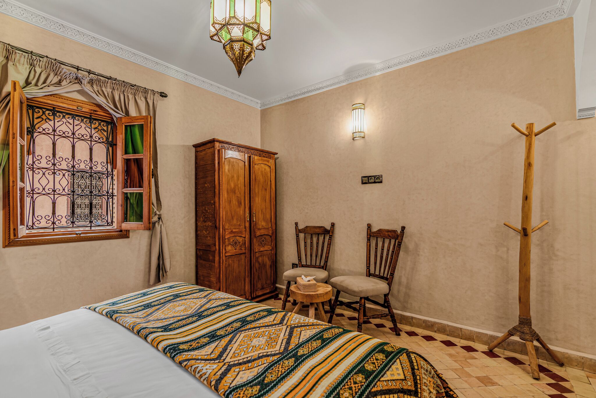 A room with a bed, two chairs, a wooden wardrobe, a coat rack, and a window with curtains.