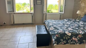 Et soveværelse med to åbne vinduer, en seng med blomstret sengetøj, en bænk og en radiator.