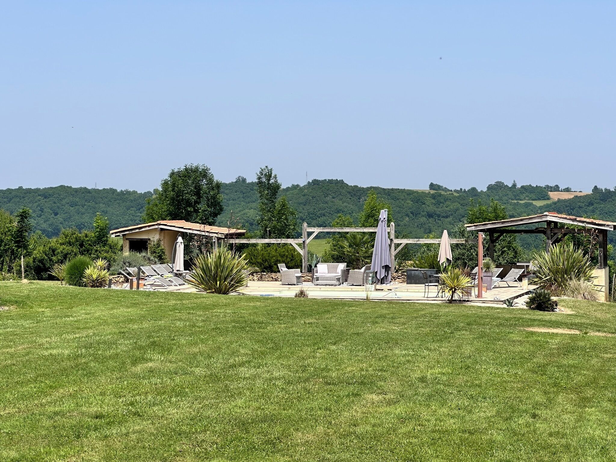 Een verzorgd buitengebied met een pergola, lounge stoelen en een klein gebouw, omringd door weelderige begroeiing en heuvels op de achtergrond.