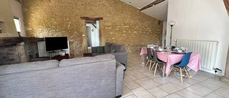 A rustic interior with exposed stone walls, a stone fireplace, a grey sofa, and a dining table set for six.