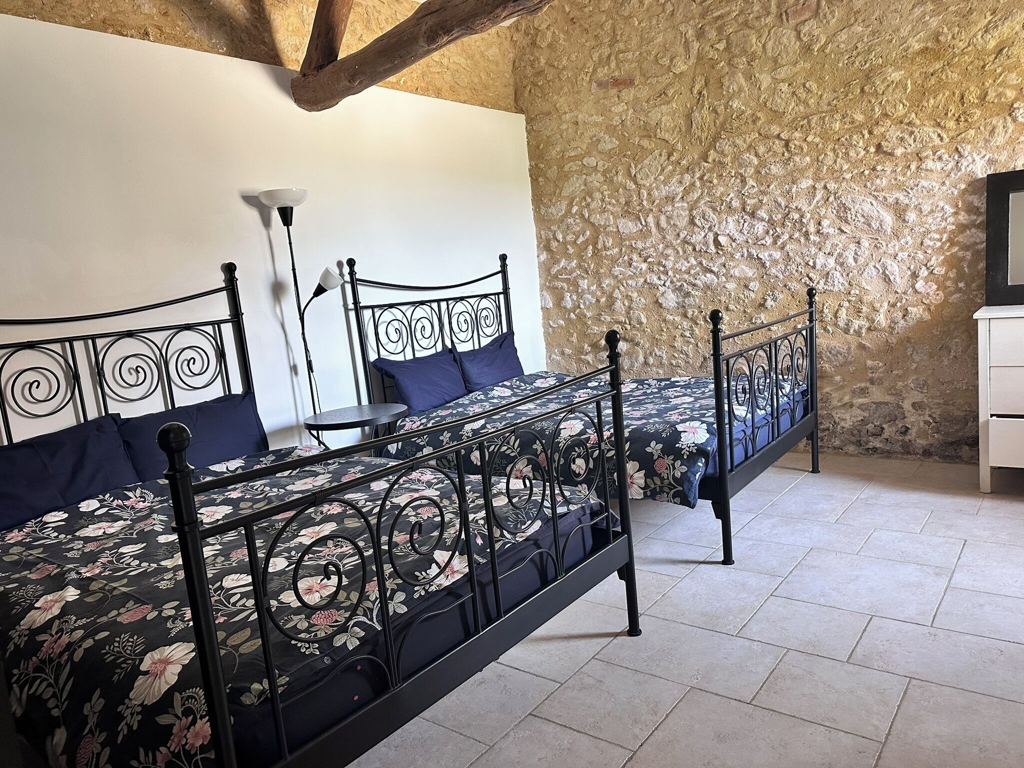 Een kamer met twee zwarte metalen bedden met bloemenbeddengoed, een klein rond tafeltje en een witte ladekast.