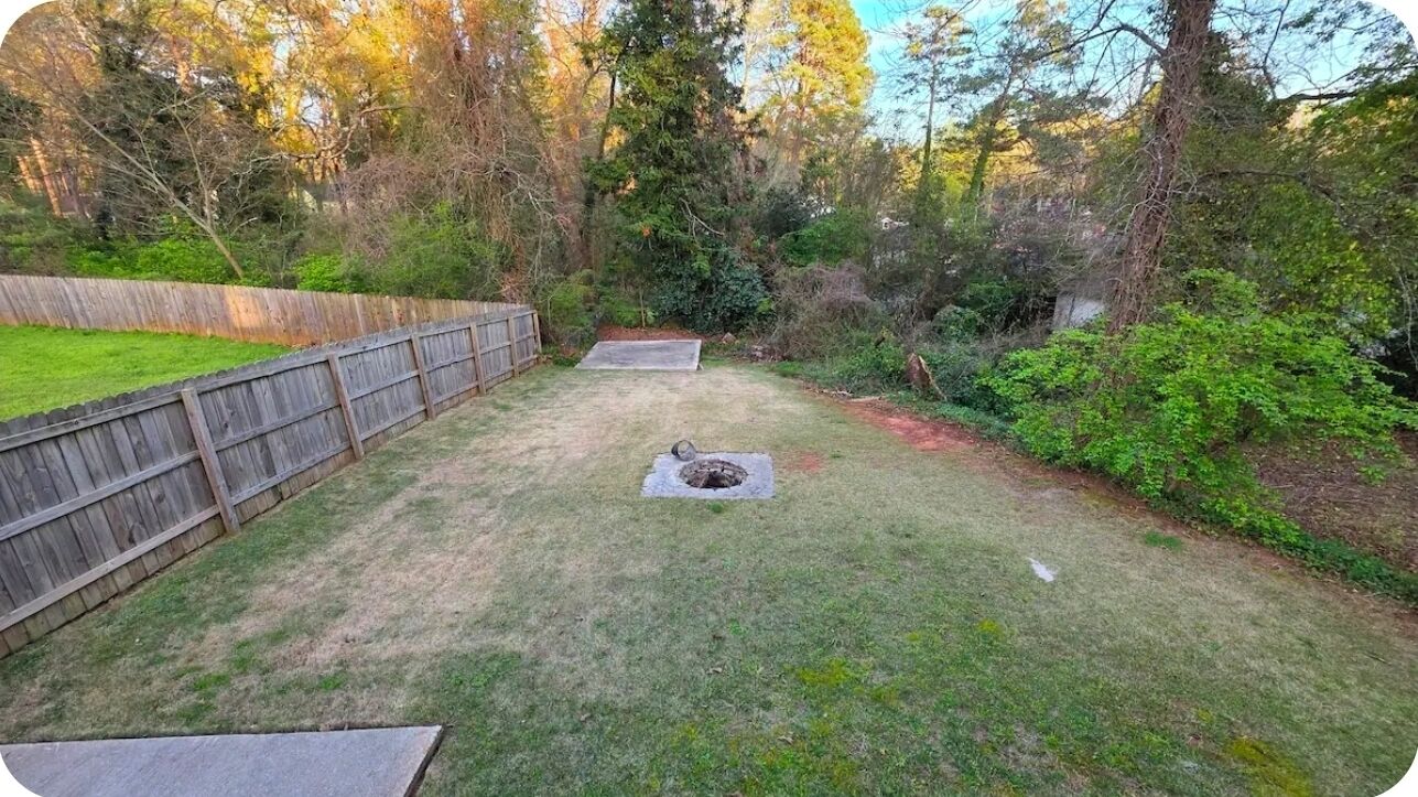 A backyard with a wooden fence, a fire pit, and a grassy area surrounded by trees.