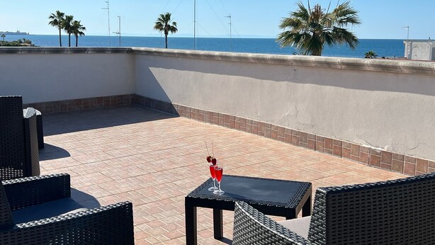 A rooftop terrace with wicker furniture, a table with a drink, and a view of the sea and palm trees. - Luca's House (Giovinazzo)