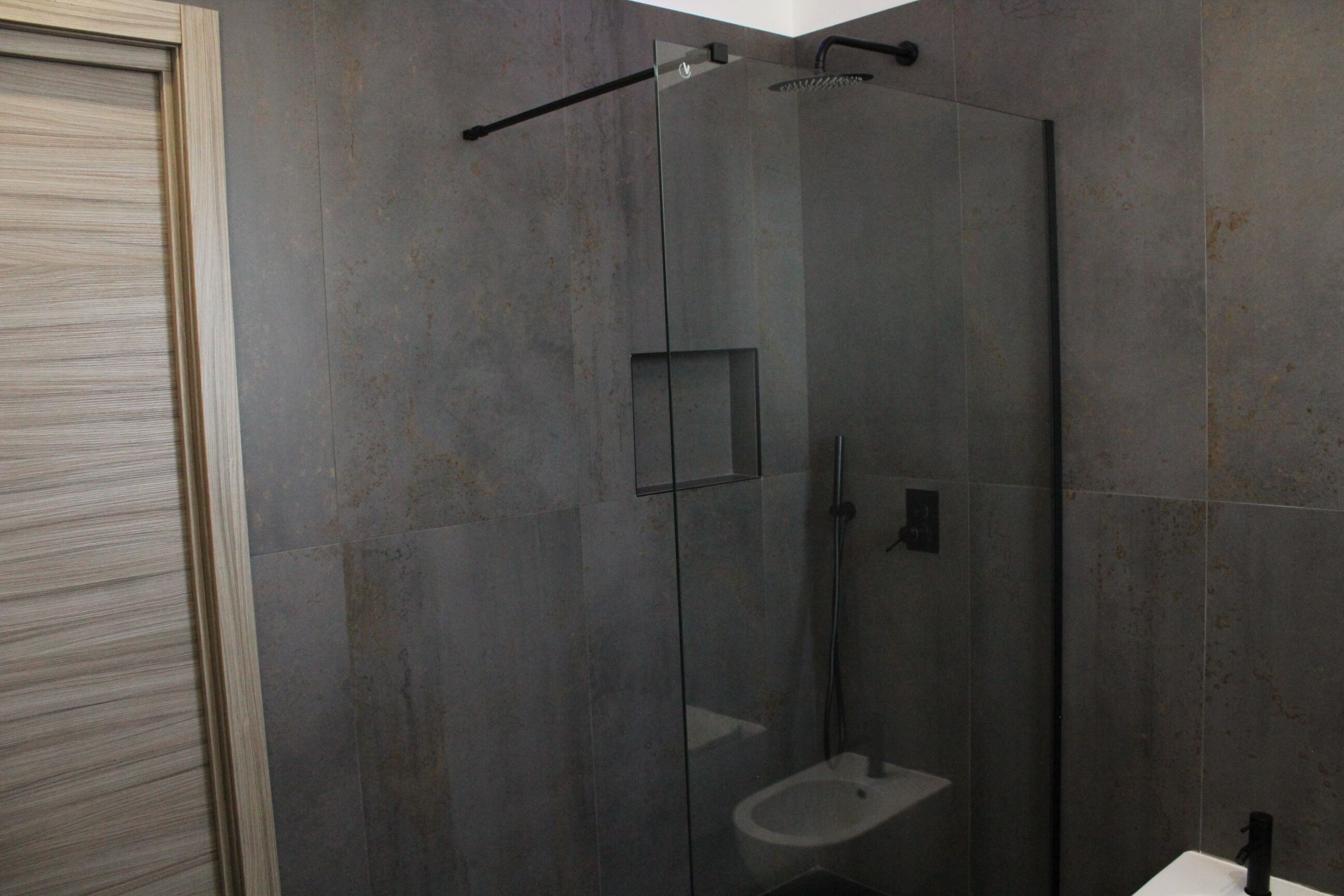 Un bagno moderno con doccia vetrata, lavandino bianco e pareti rivestite di piastrelle scure.