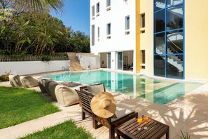 Ein moderner Poolbereich mit klarem Pool, einer Holzbank, einem Tisch mit Getränken und einem Gebäude mit großen Fenstern und einer Treppe.