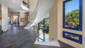 Un interior espacioso con una escalera curva, un comedor y grandes ventanales con vistas a una piscina y zonas verdes.
