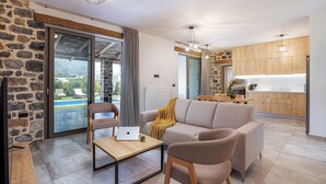 A modern living room with a sofa, a coffee table, and a laptop. There is a kitchen area with wooden cabinets and a view of the outdoors.