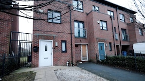A multi-story brick building with a gated entrance, a white door, and a small garden area with gravel. - Cozy Place in Salford, Sleeps 8, 4 Bedroom, 5 Beds (Salford)