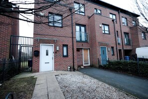 A multi-story brick building with a gated entrance, a white door, and a small garden area with gravel. - Cozy Place in Salford, Sleeps 8, 4 Bedroom, 5 Beds (Salford)