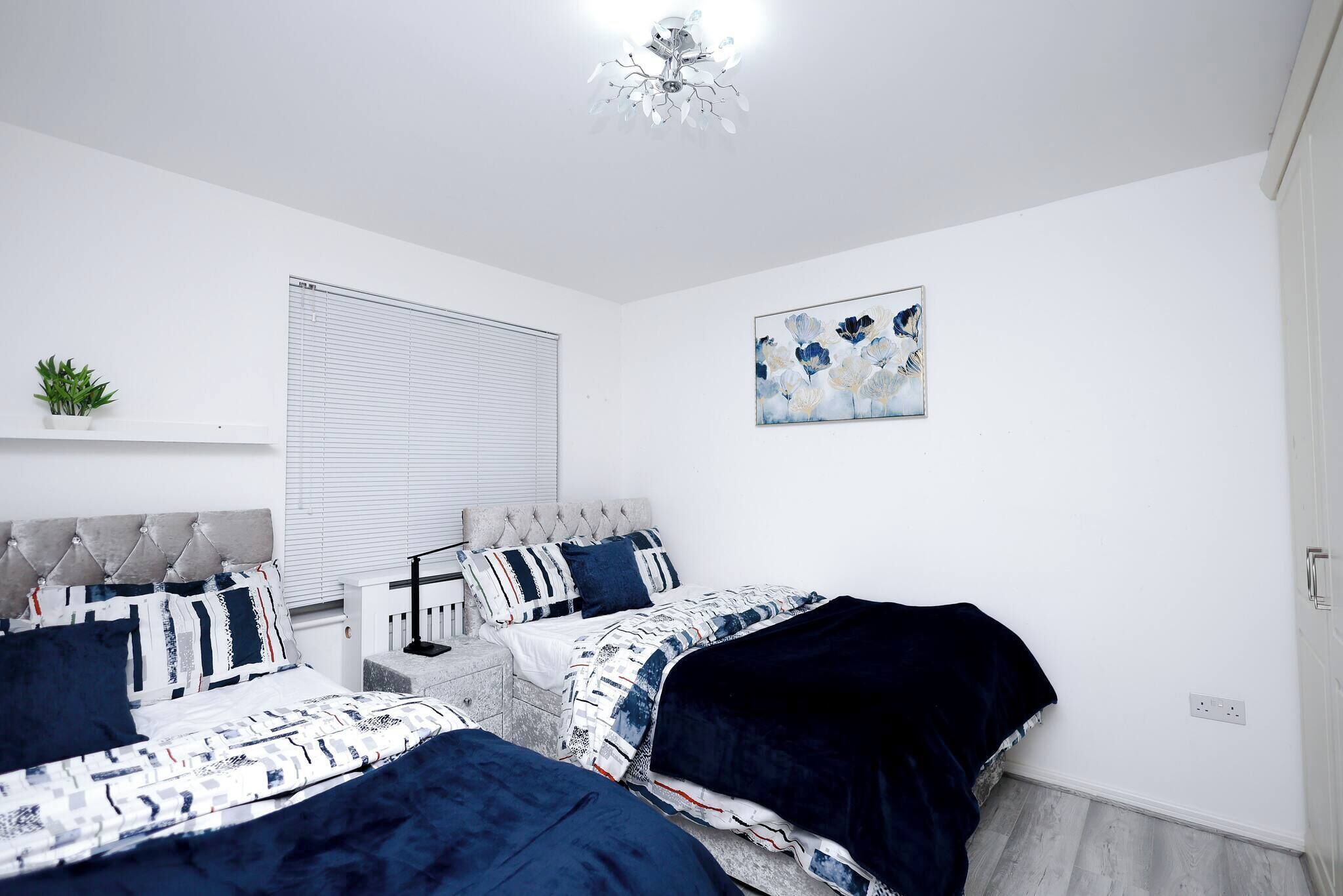 A bedroom with two beds, a window with blinds, a ceiling light, a wall-mounted plant, and a painting.