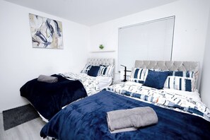 A bedroom with two beds, each with a headboard and pillows, a bedside table, a small plant on a shelf, and a window with blinds. - Cozy Place in Salford, Sleeps 8, 4 Bedroom, 5 Beds (Salford)