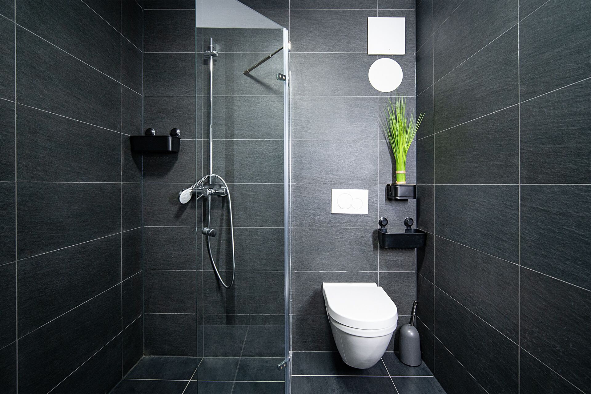 Et moderne badeværelse med mørke fliser, en bruseniche, toilet og en lille hylde med en plante.