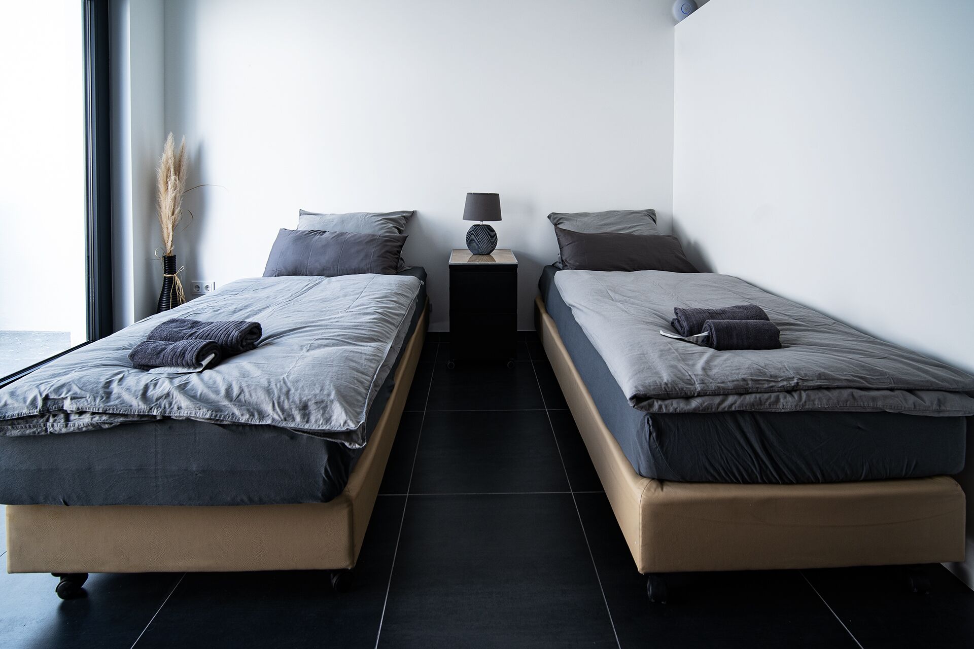 A modern bedroom with two beds, a bedside table with a lamp, and a vase with dried plants.