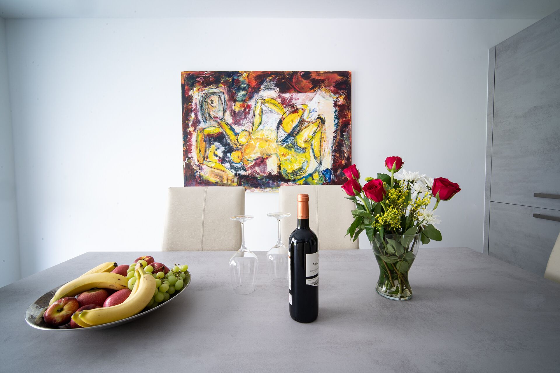 Una mesa de comedor con una botella de vino, un florero y un frutero.