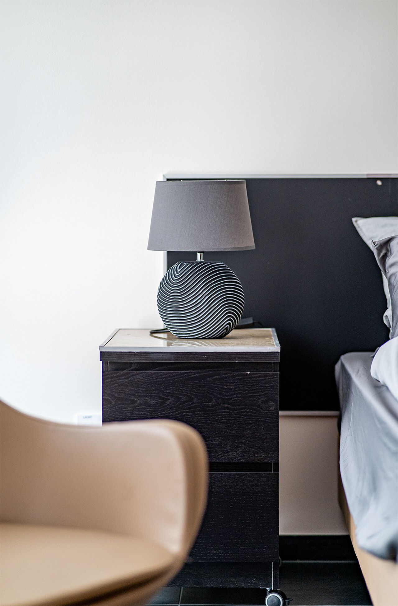 Un dormitorio moderno con una mesita de noche negra, una lámpara estampada, una cama con cabecera gris y una silla color beige.