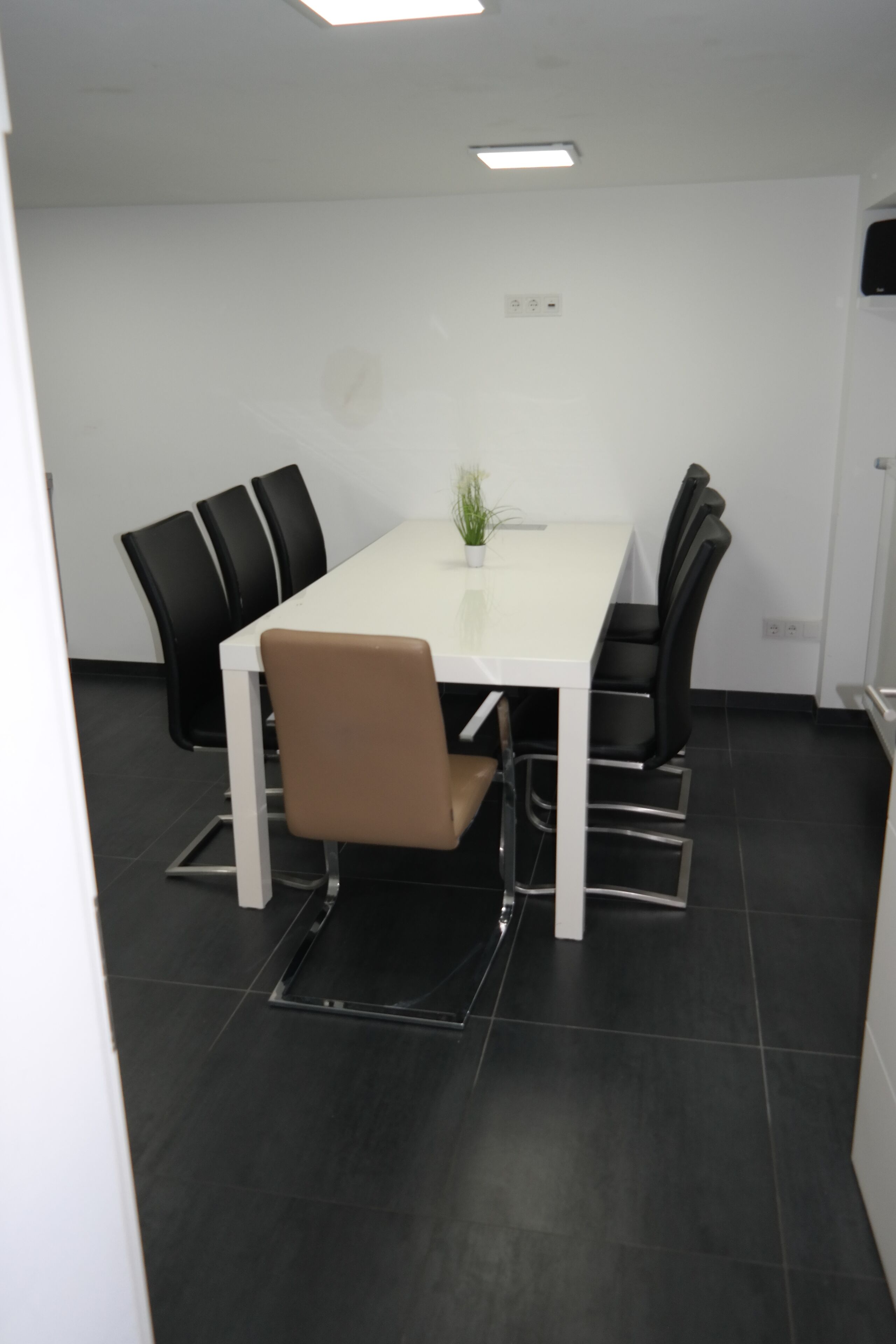 Et mødelokale med et hvidt bord, sorte og brune stole, en potteplante og et flisegulv.