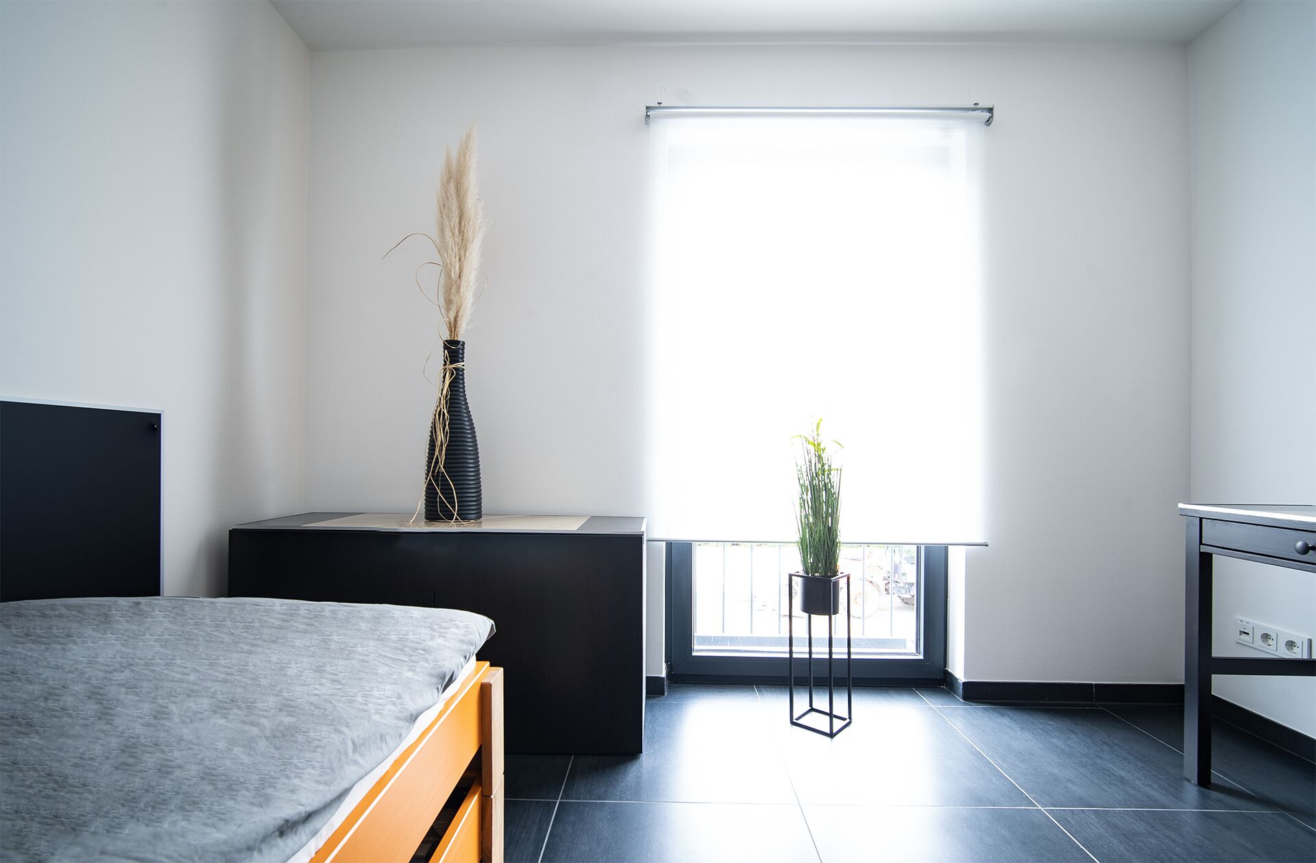Un dormitorio moderno con cama, una mesita de noche con un florero, una ventana con vistas y una pequeña planta en maceta.