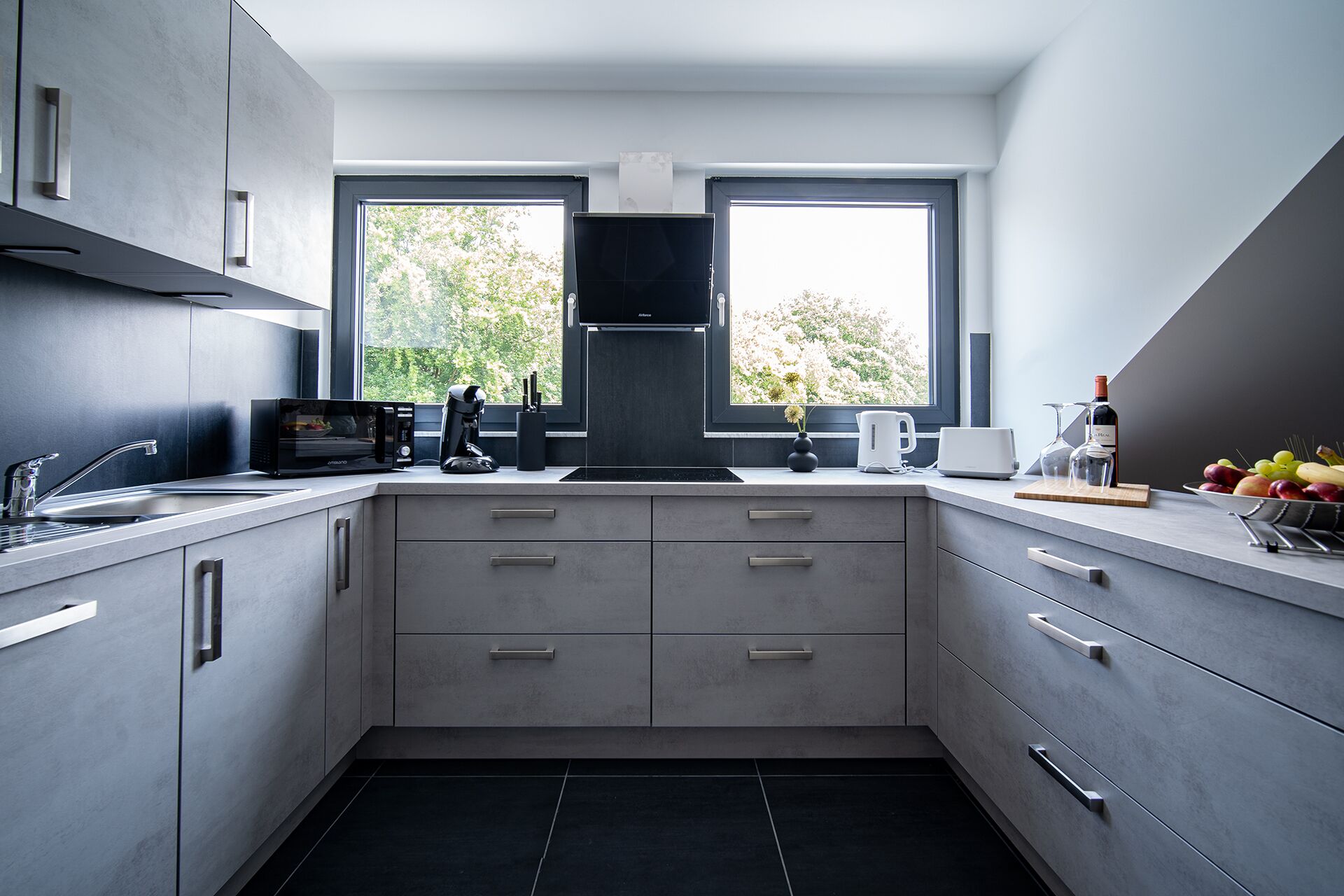 A modern kitchen with a built-in oven, microwave, and coffee maker. There are two windows providing natural light and a view of trees.