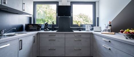 A modern kitchen with a built-in oven, microwave, and coffee maker. There are two windows providing natural light and a view of trees.