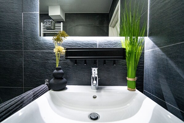 A bathroom with a white sink, dark tiled walls, a towel rack, and a plant.