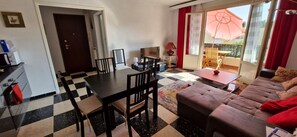 A living room with a checkered floor, a sofa, a dining table with chairs, a TV, and a balcony with a red umbrella. - LE FLOREAL Apartment 75 m² - 2 Bedrooms - Sleeps 4 (Bandol)