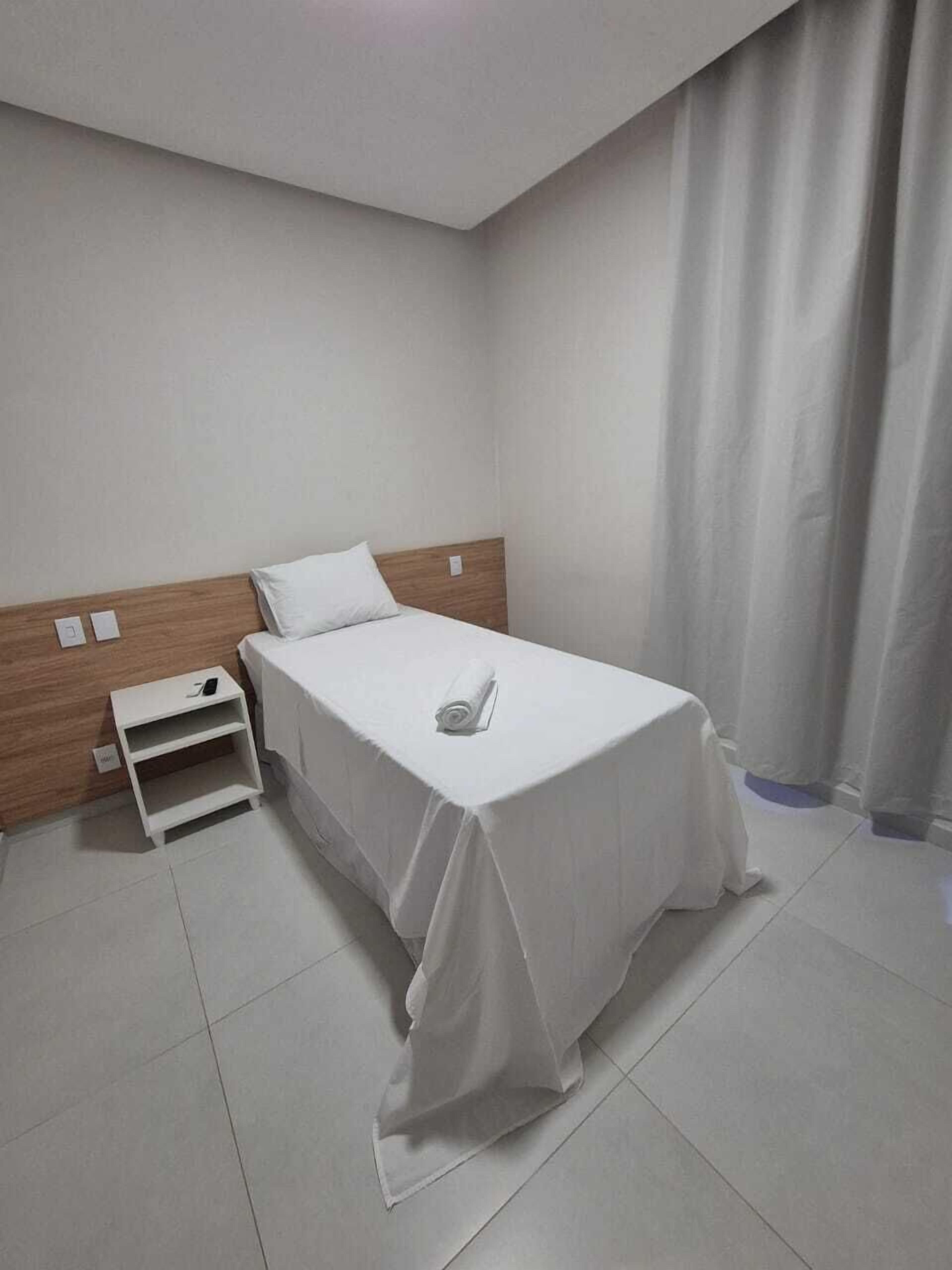 Una cama bien hecha con sábana blanca y una toalla enrollada, cabecera de madera, mesita de noche blanca con un teléfono y pared blanca.
