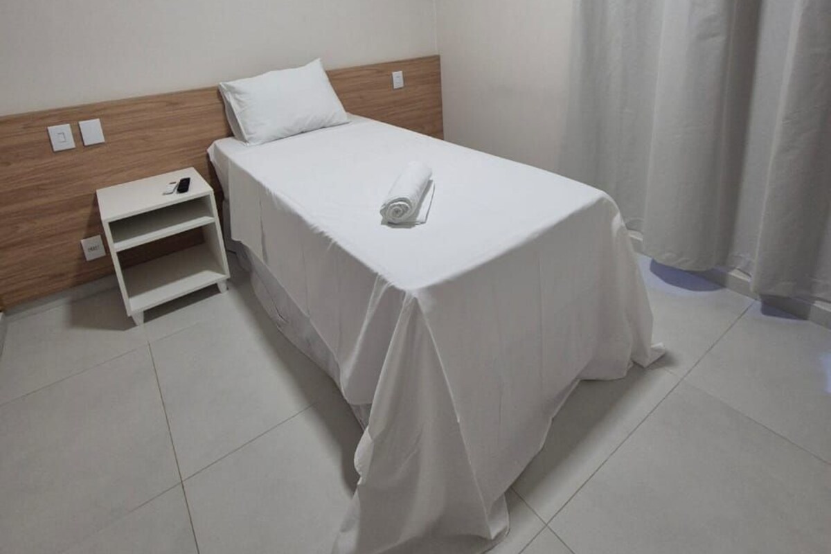 A neatly made bed with a white sheet and a rolled towel, a wooden headboard, a white nightstand with a phone, and a white wall.
