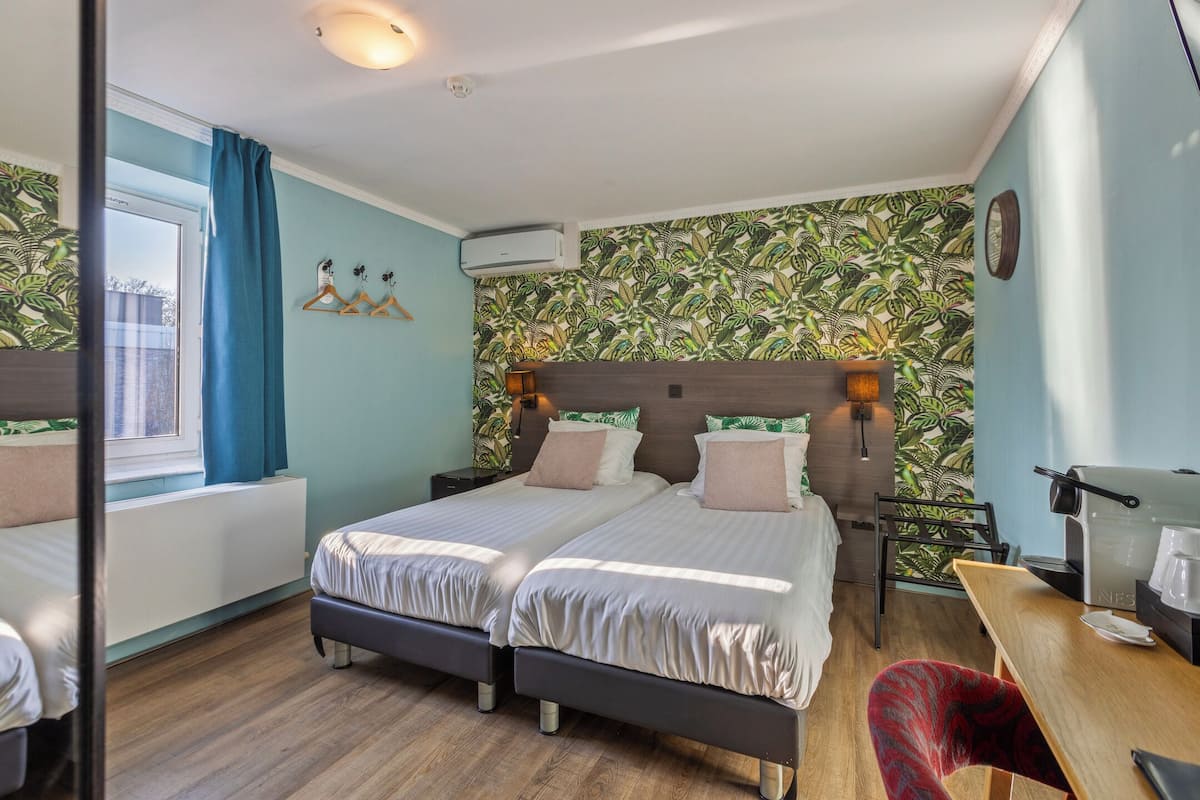 Une chambre avec deux lits, un bureau en bois, une chaise, une fenêtre avec des rideaux et un mur orné d’un motif végétal.