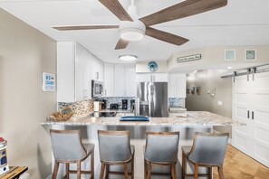A kitchen with white cabinets, a stainless steel refrigerator, a ceiling fan, and a bar area with three chairs. - Ponte Vista 206 – Waterfront Comfort & Sunset Views (Ocean City)