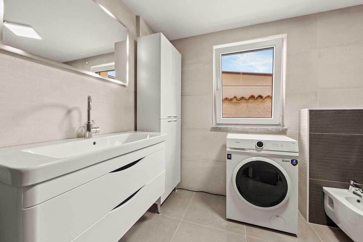 Une salle de bain moderne avec un grand lavabo, un miroir, un lave-linge et une fenêtre donnant sur un paysage désertique.
