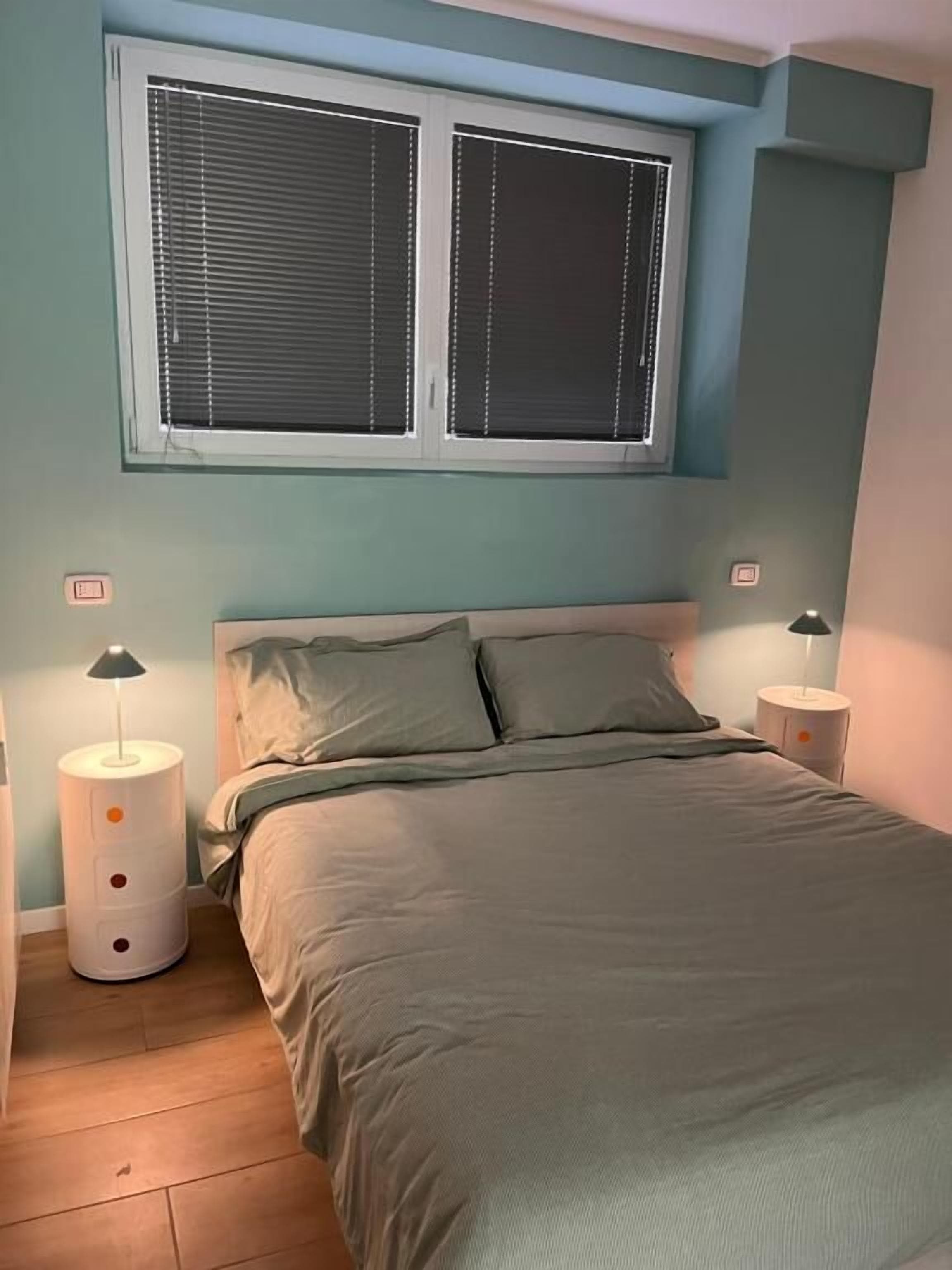 Een slaapkamer met een bed, twee nachtkastjes, een raam met zonwering en een wandthermostaat.
