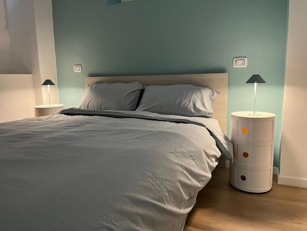 A neatly made bed with a grey comforter and pillows, two bedside tables with lamps, and a wall-mounted thermostat.