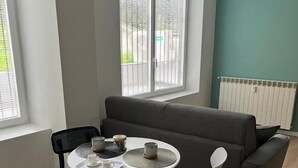 A modern living room with a grey sofa, a round dining table with chairs, and a window with blinds. - B&B Malnati 3 Urban Living Milanosansiro (Milan)