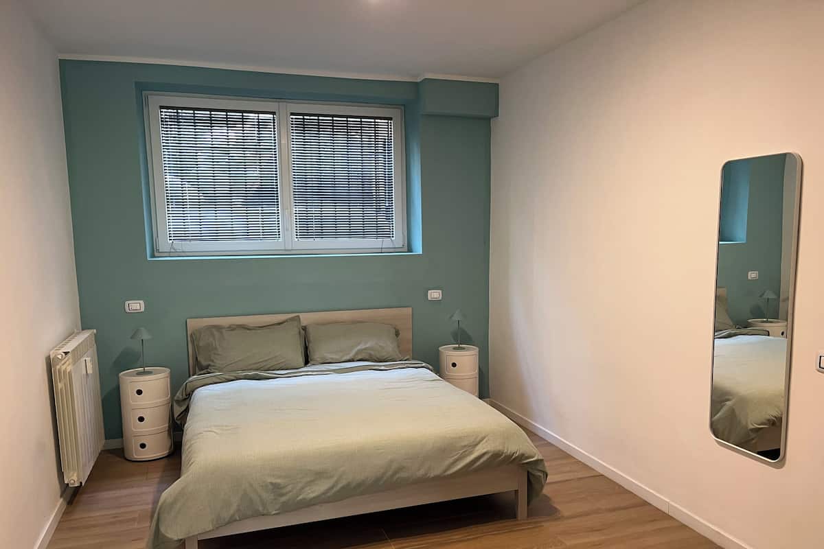 Une chambre à coucher comprenant un lit, des tables de chevet, un radiateur, une fenêtre avec des stores et un miroir au mur.
