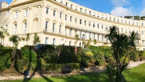A large, classical building with symmetrical architecture, featuring a central entrance, multiple windows, and a well-maintained garden with palm trees and a pathway. - Meadfoot Bay Apartment at Hesketh Crescent (Torquay)