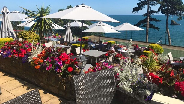 An outdoor seating area with tables and chairs, surrounded by colorful flowers and white umbrellas, overlooking a body of water. - Meadfoot Bay Apartment at Hesketh Crescent (Torquay)