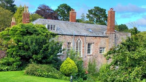 A stone house with multiple chimneys, a prominent window, and lush greenery surrounding it. - Lower Stable - The Stables Apartments at Meadfoot Bay (Torquay)