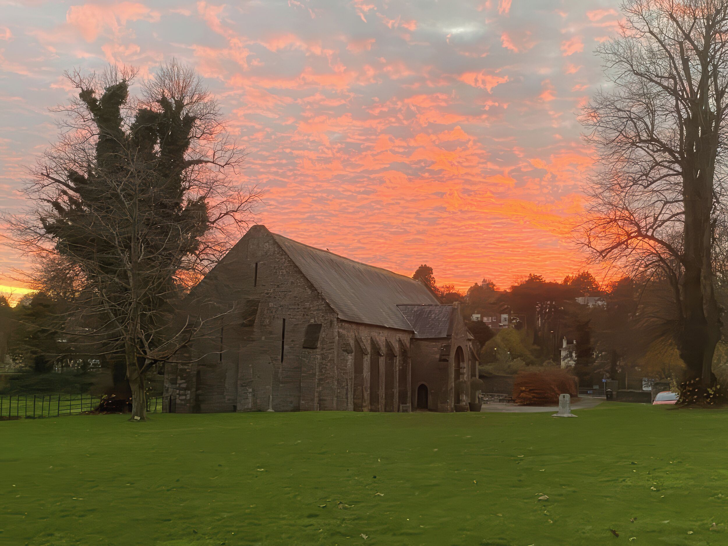 A stone building with a triangular roof, surrounded by a grassy field and trees, under a sunset sky.